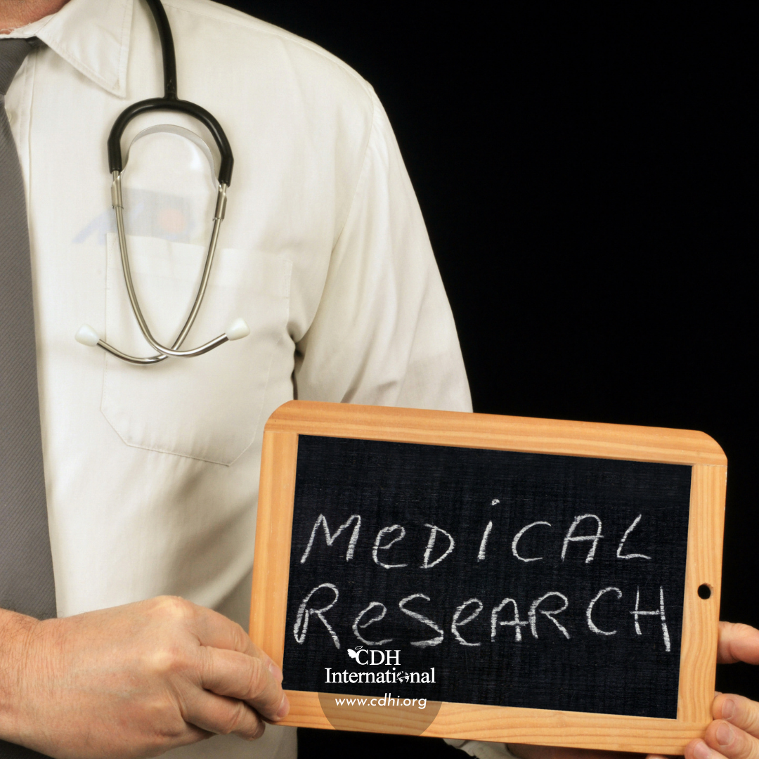 Doctor in a white coat with a stethoscope holds a chalkboard that reads 'Medical Research' (CDH International logo visible).