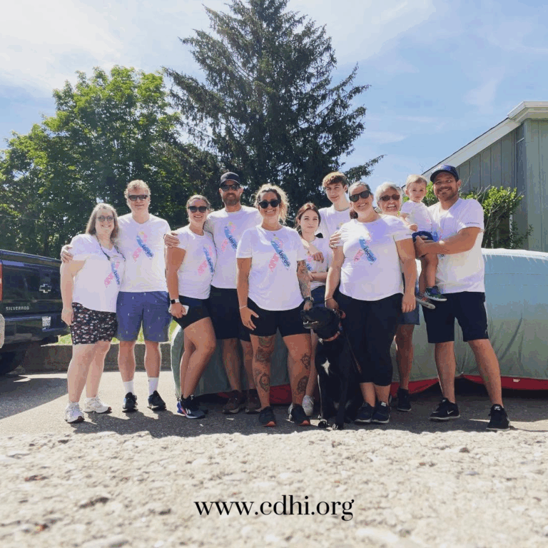 Group of people in matching white shirts posing outdoors for a charity event, with a black dog at the center and 'www.cdhi.org' visible at the bottom.