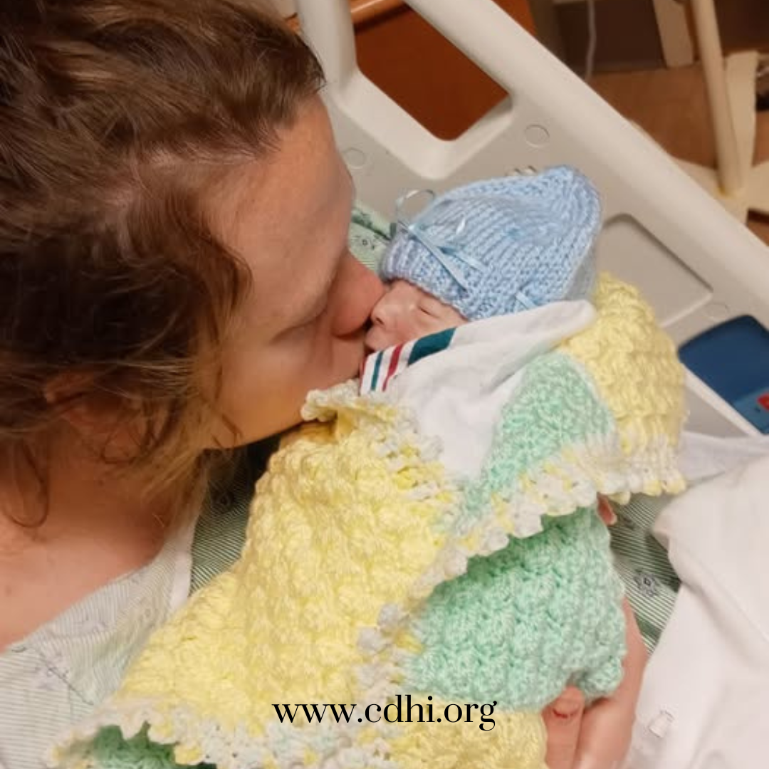 Mother kissing her newborn baby who is wrapped in a pastel crochet blanket and wearing a blue knit hat in a hospital bed.
