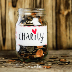 Glass jar filled with coins on a wooden surface, label reads 'CHARITY' with a red heart.