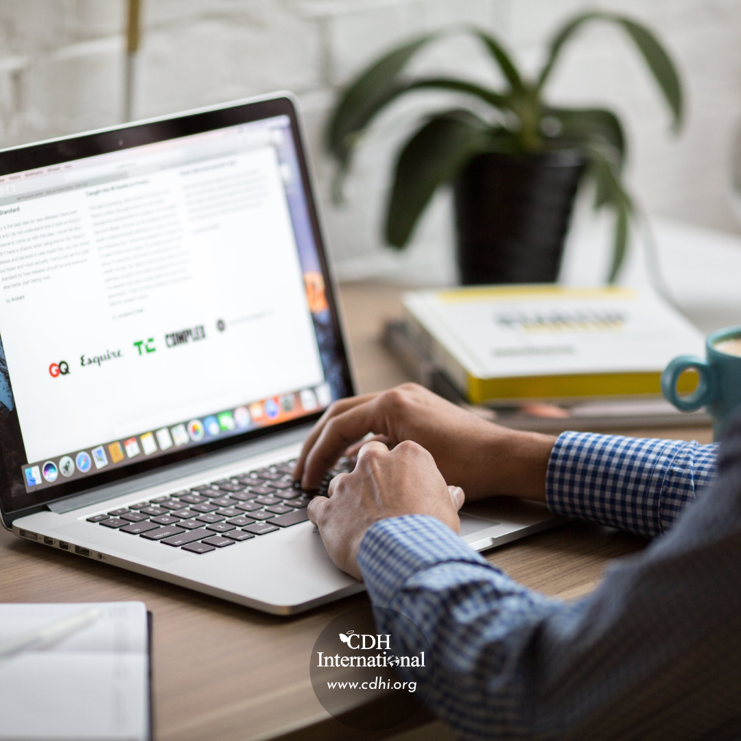 Person typing on a laptop at a wooden desk, with a plant and books in the background and a watermark for CDH International on the screen.