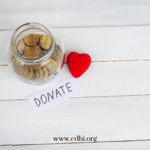 Glass jar filled with coins beside a red plush heart and a 'DONATE' note on a white wooden surface, www.cdhi.org.