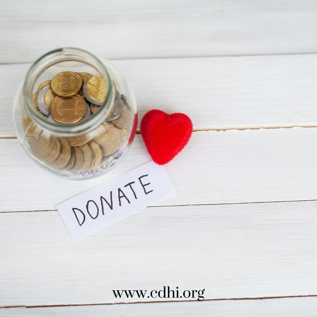 Glass jar filled with coins beside a red plush heart and a 'DONATE' note on a white wooden surface, www.cdhi.org.