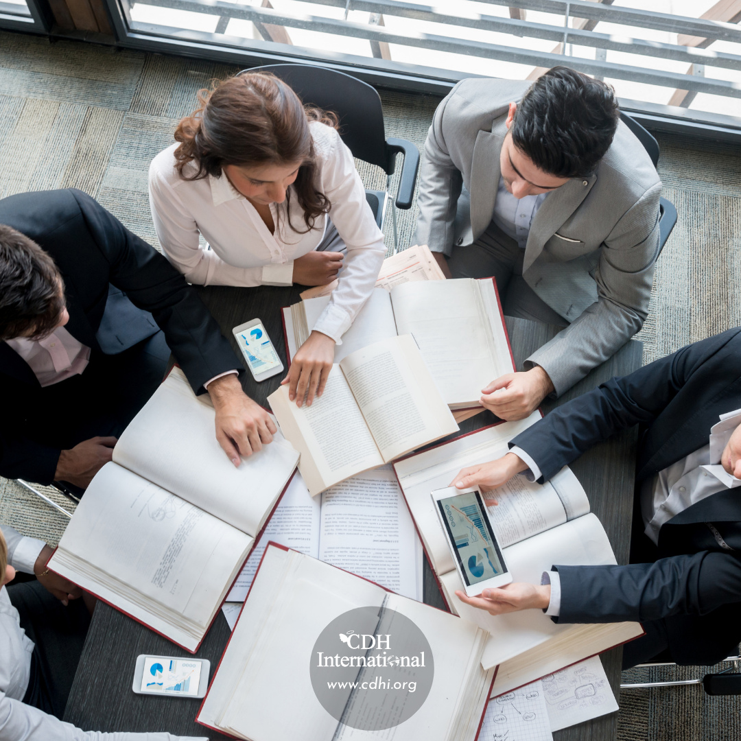 Top-down view of five professionals around a table with open books and smartphones, collaborating on study material; CDH International logo visible.