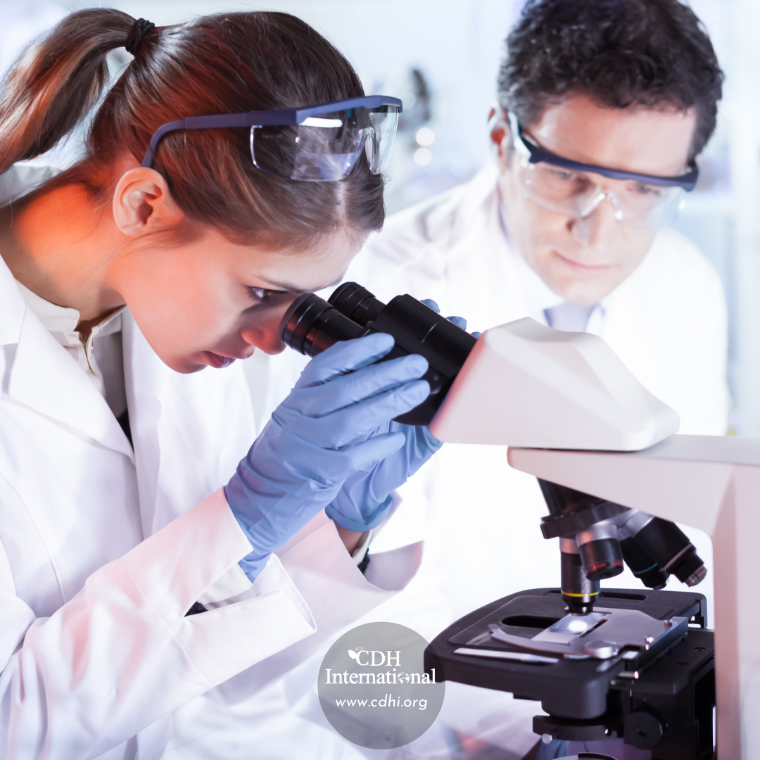 Two scientists in white lab coats and goggles examine samples under a compound microscope to study them closely.