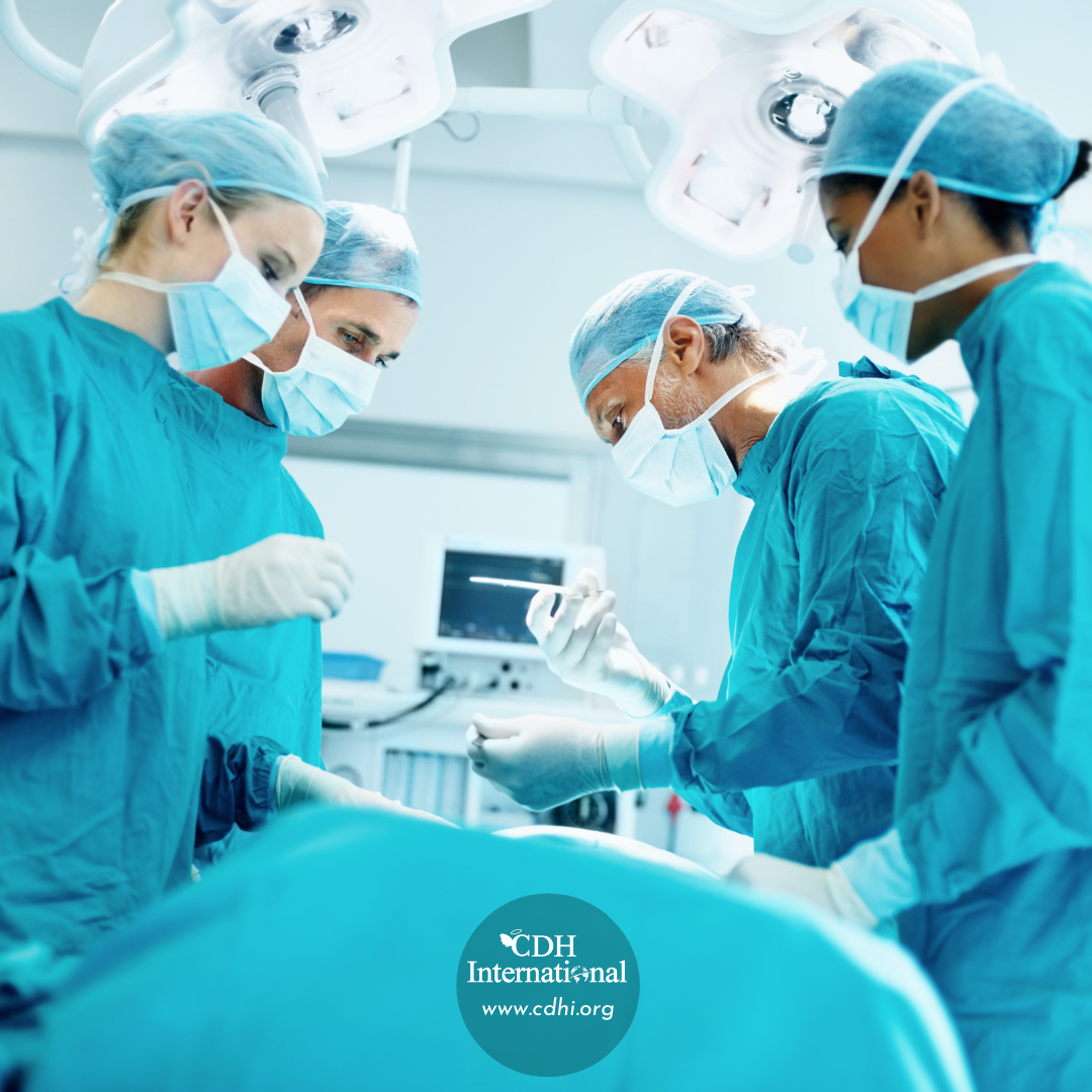 Surgeons in blue scrubs and masks perform an operation under bright overhead lights in an operating room, with a patient out of view and a monitor in the background, CDH International logo shown at bottom.